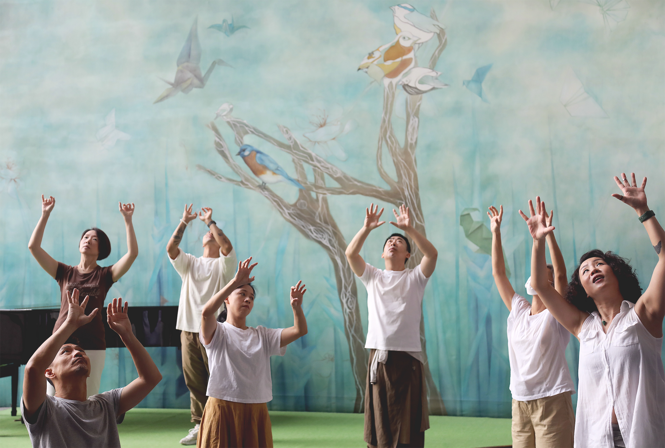 In front of a mural with light blue background with a tree trunk, a few birds and some paper cranes, seven dancers reaching their hands towards the sky, heads looking up.