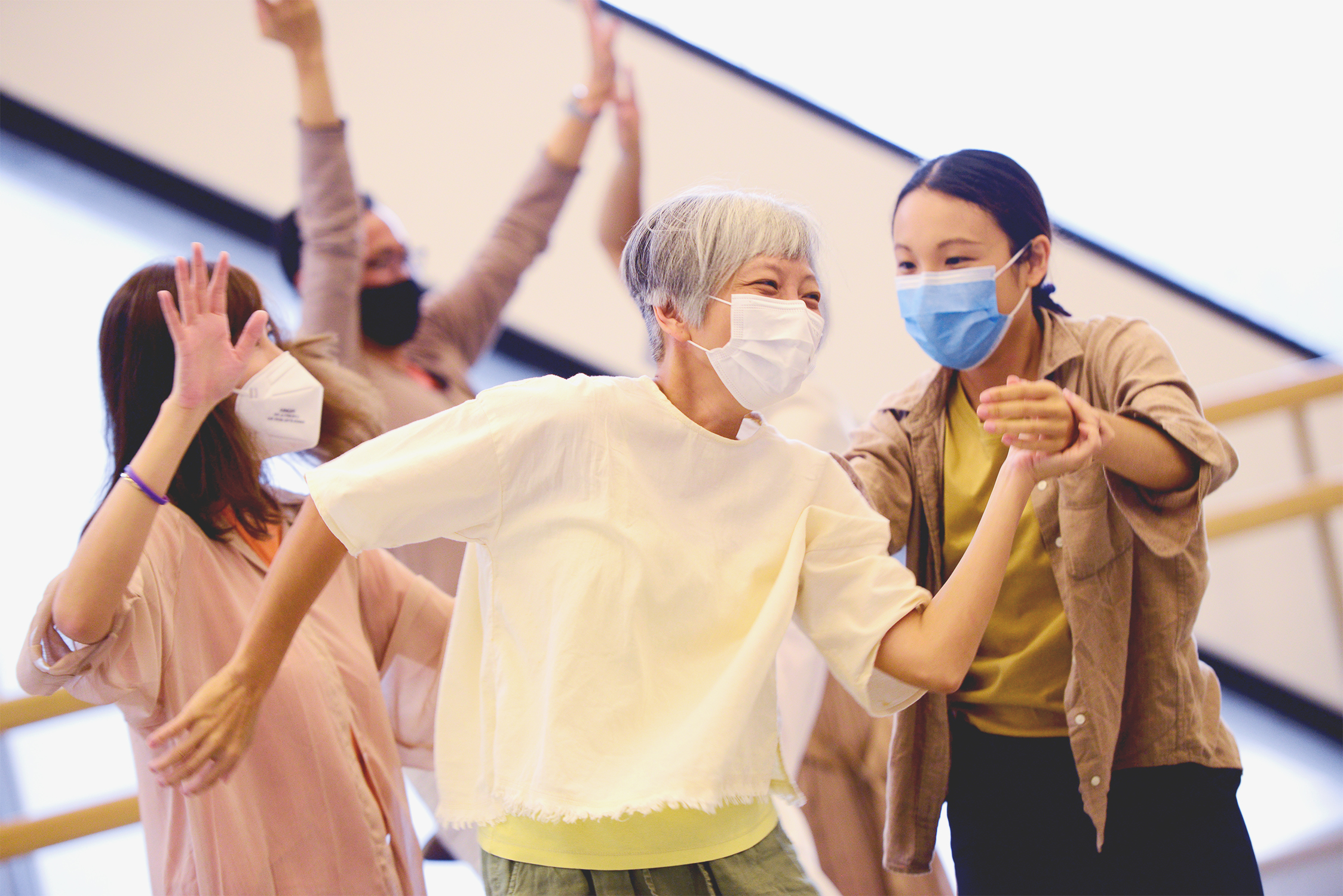 Five dancers enjoying inside a dance studio, at the front, there are two female dancers, an elder one and a younger one, both dancing together, the elder dancer’s left hand in front of her own body, holding the left wrist of the younger dancer.